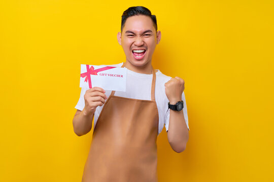 Excited young Asian man 20s barista employee in brown apron working in coffee shop, holding a gift store coupon voucher card, celebrating success on yellow background. Small business startup concept