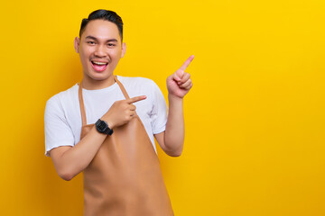 smiling young Asian man 20s barista barman employee wearing brown apron working in coffee shop, pointing finger on advertising area on yellow background. Small business startup concept