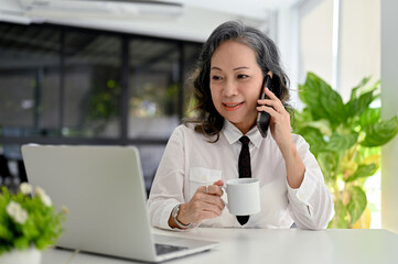 Professional Asian aged businesswoman working in office, talking on the phone and sipping coffee.