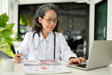 Fototapeta premium Professional Asian aged female doctor working in the doctor's office, using laptop.