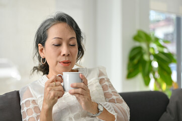 Peaceful and relaxed elderly Asian woman sipping morning coffee while chilling in living room.