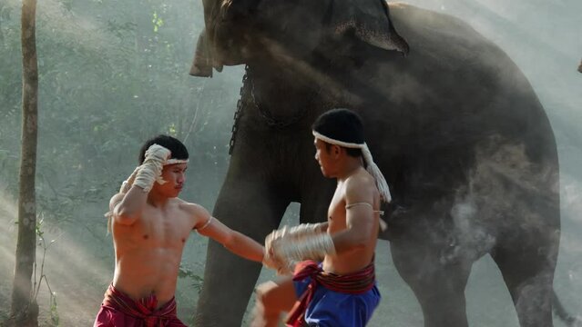 Two Young males wearing Thai tradition short, head and hands wrapped in twisted hemp rope and showing beautiful fighting art of Muay Thai, Blurred elephants and spreading white mist in background