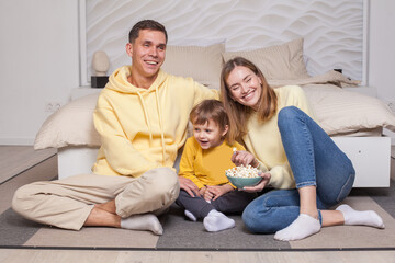Cute laughing smiling parents with child son sitting by the bed and eating popcorn