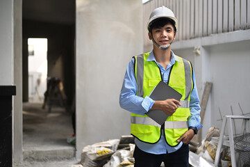 Portrait of architect man in safety helmet and reflective vest standing front of construction site and smiling to camera
