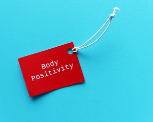 Red paper tag on blue background with text BODY POSITIVITY, the movement to  shift unrealistic feminine beauty standards into a more whole-bodied, realistic approach, all bodies are beautiful