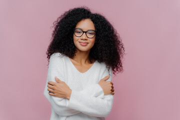 Pleasant looking millennial Afro American woman keeps hands crossed over chest, has curly bushy...