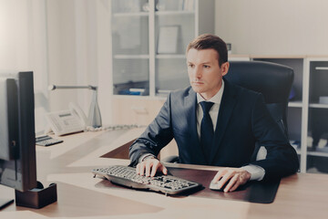 Fototapeta na wymiar Serious concentrated businessman sits in front of computer at work place, keyboards information, wears suit, prepares business project for next year, poses in spacious cabinet. People and finances