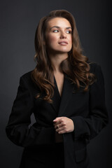 Fashion, business and make-up concept. Portrait of beautiful brunette woman with black suit and big wavy hair in dark studio background. Model standing in grace pose and looking aside camera