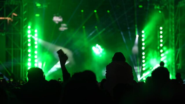 Crowd of fans are cheering at open-air music concert on illuminated stage with neon colorful lights, back-view