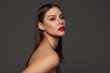 Portrait of female fashion model with naked shoulders and long hair looking at camera over dark grey background. Beautiful woman with trendy make-up, red lipstick and well-kept skin. - obrazy, fototapety, plakaty
