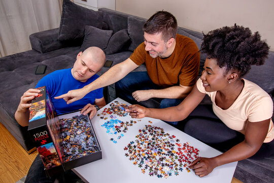 Friends playing with jigsaw puzzle at home, on a white wooden table. Putting things together and solving problems. Fun and diversity in friendship. - Powered by Adobe