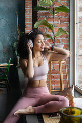 Beautiful young American woman enjoying listen music with big white headphones on windowsill after sport training. Concept of music in daily youth life. Vertical
