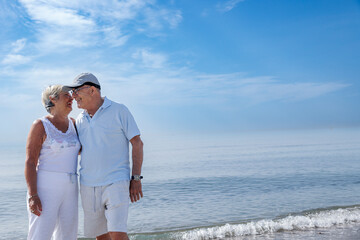 coppia di anziani coniugi si abbraccia affettuosamente vicino alla riva in una spiaggia  - obrazy, fototapety, plakaty