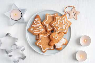 Top view of homemade spiced baked gingerbread cookies or crunchy biscuits cooked with ginger spice decorated with sweet sugar icing served on plate on white wooden background with glowing candles