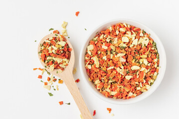 Mixed pieces of dried carrots, celery root and greens in ceramic bowl and wooden spoon on white background.  Vegetable culinary condiment
