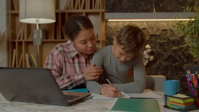 Smart adorable preadolescent multiracial boy with caring charming black mother doing homework for elementary school, writing task into textbook while studying together in domestic room.