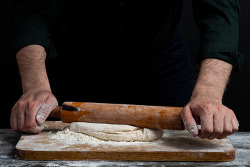 Male hands with wooden rolling pin rolls out dough. Close Up view of baker's male hands making...