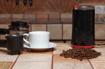 Preparing coffee: coffee beans, electric coffee grinder, coffee maker horn and white cup and saucer on a tiled kitchen table