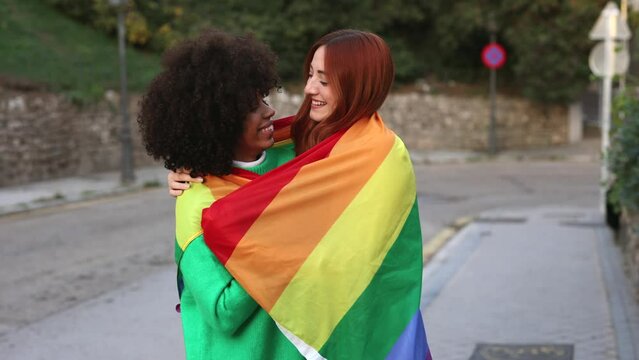 Multiracial lesbian couple look at each other with love hugging with lgbt freedom rainbow flag, diverse people love each other