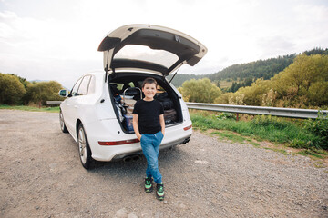 Child stands next to the full trunk of an SUV. Child comes with us on vacation and it is driven by car in the mountains. Full suitcases in the trunk of a car