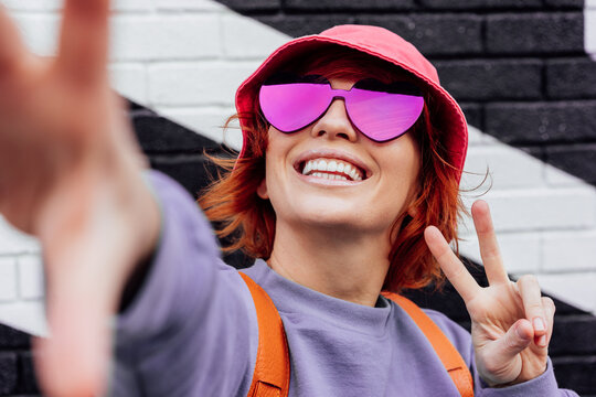 Close-up excited redhead woman in magenta heart-shaped sunglasses smiling and showing V finger sign while taking a selfie photo outdoors. Color of the 2023 year. Emotional hipster fashion women