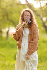 young woman plucks wild blue flowers in the green park on summer sunny day. girl with curly hair in dress makes bouquet of beautiful flowers. vertical photo