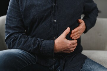 Unhappy senior man stomach ache, man presses hand to stomach from unbearable pain, mature man with stomach pain feeling unwell sitting in living room
