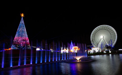 Christmas decoration set consisting of a Christmas tree, water with lights and a giant Ferris wheel. Night photo of Christmas lights. - obrazy, fototapety, plakaty