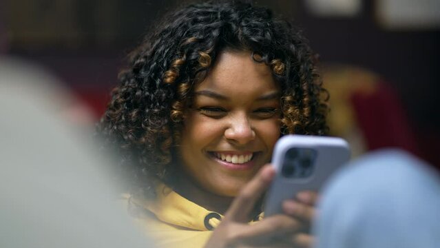 One happy young black woman laughing while holding smartphone on couch. African American 20s girl staring at cellphone in the evening