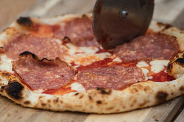 Cutting hot pizza with tomato sauce, mozzarella cheese and salami meat with a cutter on a wooden board, with smoke, close up