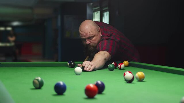 A big bald brutal bearded man plays billiards on a green table, takes aim and hits a white ball with a cue. Pool balls with numbers on a green-clothed pool table, sports game of billiards