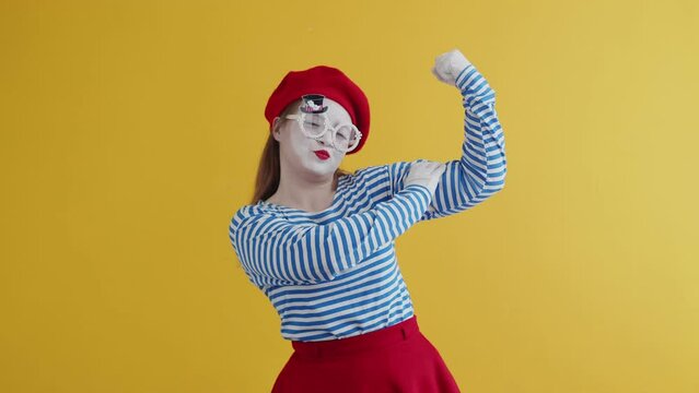 A mime girl in a striped and bright suit smiles and jokingly points to her bicep, toched it. Pantomime plays out a performance on a yellow background