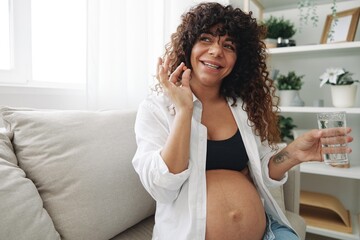 Fototapeta na wymiar Pregnant woman smile blogger takes vitamins and medicines pills with a glass of water close-up sitting on the sofa at home freelancer in the last month of pregnancy lifestyle before childbirth