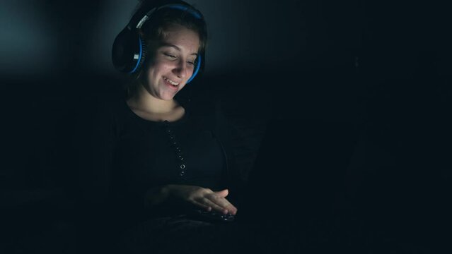 Pretty girl talking by video call wearing earphones and using laptop in dark room with no light in night time. Teenager during web cam communication