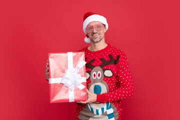 smiling man in winter sweater and santa claus hat. xmas guy with present box on red background.
