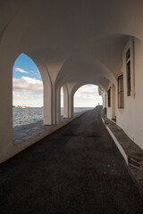 Tunel blanco con arcos por donde se ve el mar Mediterráneo con pequeñas casas al otro lado del camino.