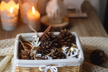 Basket with pine cones, Christmas ornaments and seasonal spices. Soft blanket, and lit candles in the background. Selective focus.