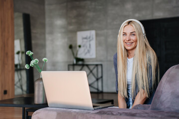 Excited blonde young woman in headphones smiles sits on couch with laptop makes looks at camera toothy smiles in adorable mood. Swedish female student girl after remote lesson via internet. Success.
