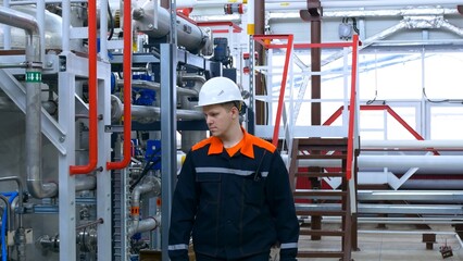 An industrial engineer in overalls and a white helmet with a blueprint in his hands stands near the technological equipment with a pipeline and pressure and temperature sensors. Factory work.