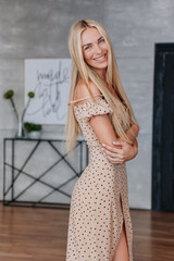 Vertical portrait of young blonde caucasian woman in beige dress looks at camera toothy smiling at home. Pretty Swedish girl with long loose hair satisfied by health and skincare products. Beauty