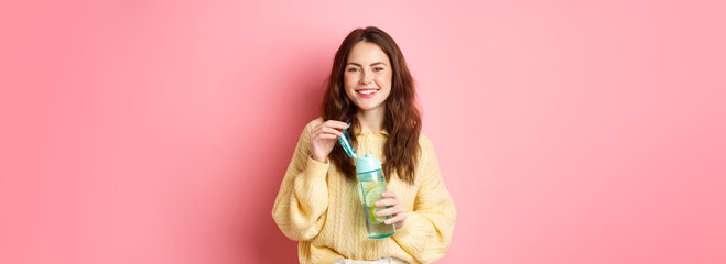 Young attractive and healthy woman smiling at camera, open water bottle with lemon, drinking sport...
