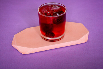 Close up of a bramble cocktail in a tumbler glass, on a peach concrete platform flatlay.