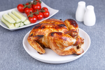 Chicken baked whole to a delicious crust. A traditional dish. In the background are cucumbers and cherry tomatoes on a white plate. Close-up, selective focus, gray background.