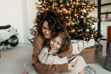 Two adorable sisters wearing sweaters sitting in front of Christmas tree. Family portrait of charming young mother and cute lovely daughter 