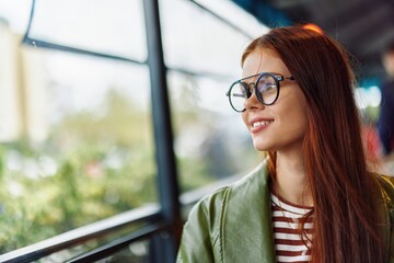 Portrait of a woman with red hair with glasses sitting in a cafe in the city by the window smile with teeth, female freelance blogger close-up