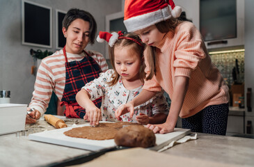 Cute little girls in red Santa hats with mother making homemade dough Christmas gingerbread cookies using cookie cutters together in home kitchen. Happy family holidays preparation, childhood concept.