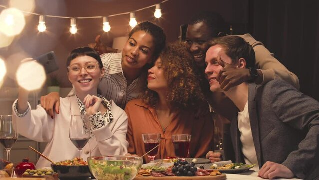Five young fancy multiethnic friends taking selfie portrait on smartphone while having festive dinner party in apartment