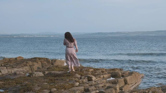 Woman in a dress walks along a rocky seashore. Firth Of Clyde, Troon, Scotland