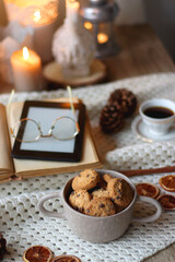 Cup of tea, books, tablet, bowl of cookies, various spices, pine cones and lit candles. Hygge at home. Selective focus.