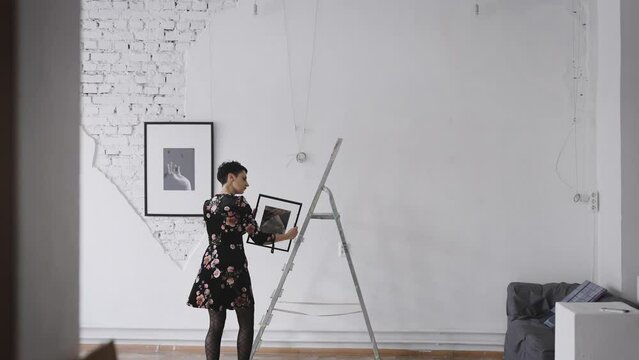 A woman gallery owner hangs new paintings and collages on the wall of the exhibition hall. Gallery with an exhibition of contemporary art.